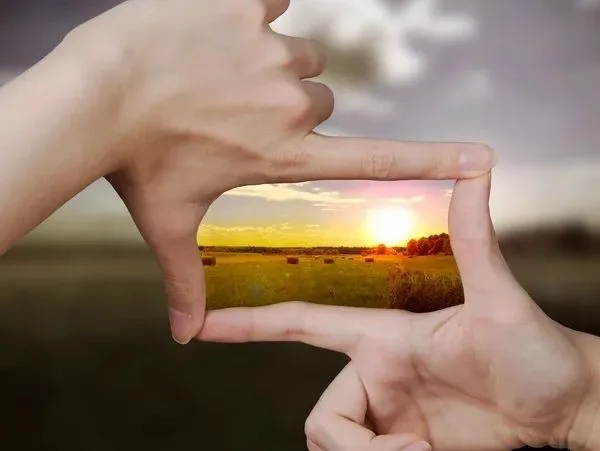 Hands framing a sunset landscape in a field with hay bales.